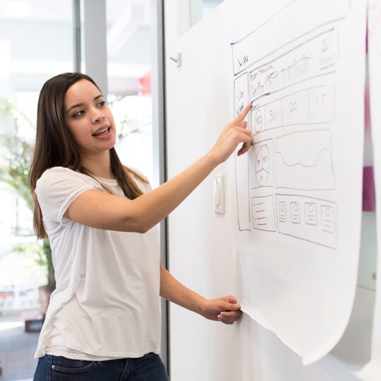 A young woman pointing at a design on a whiteboard.